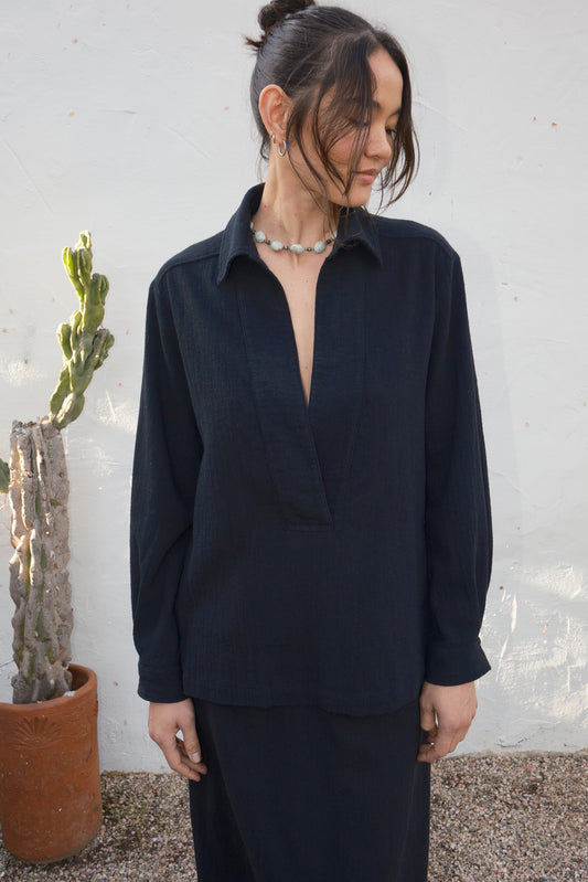 Woman wearing a black outfit standing next to a potted plant against a textured wall.