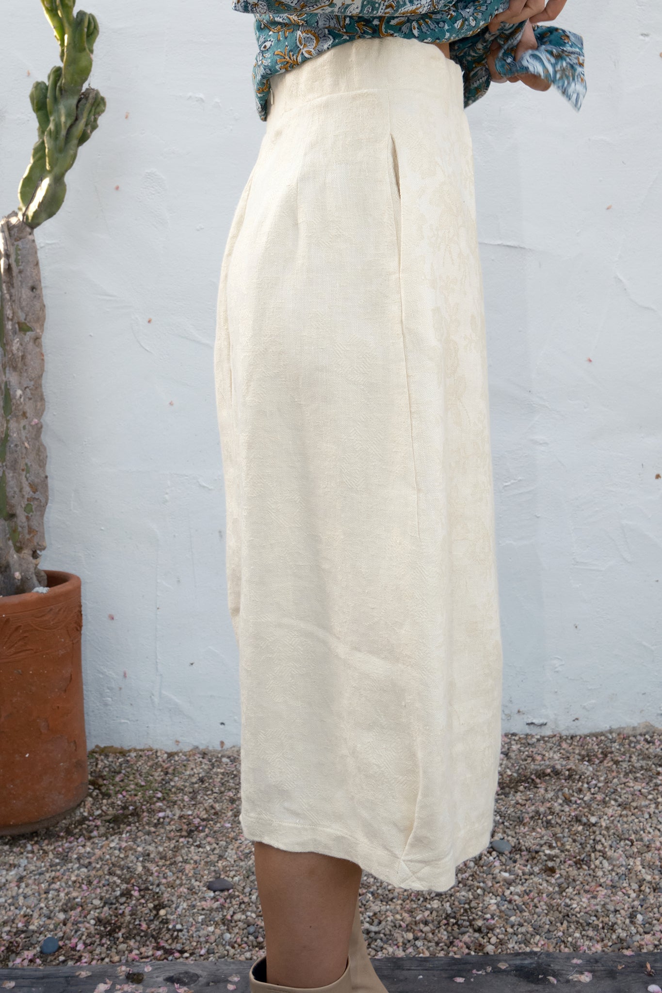 Person wearing a beige skirt standing next to a cactus plant against a white wall.