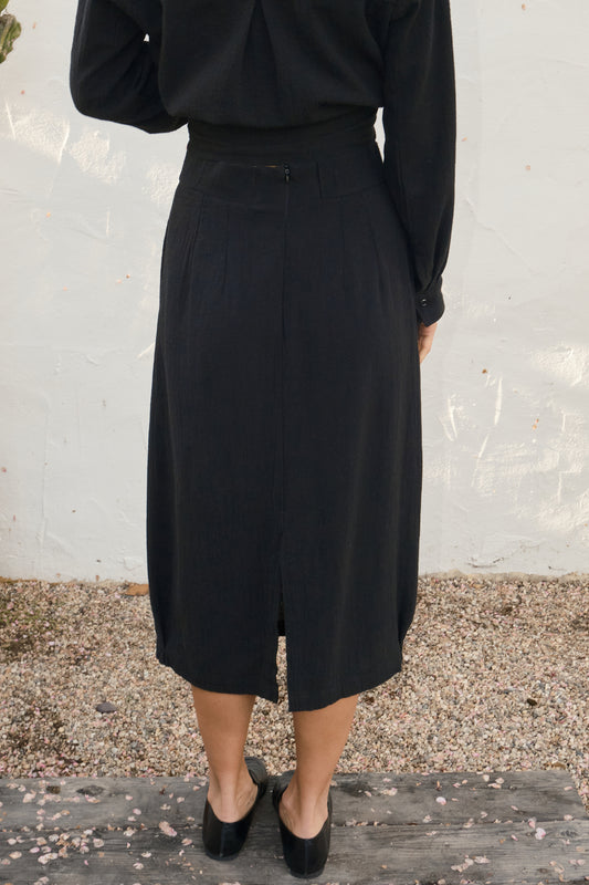 Person wearing a black outfit with a skirt against a white wall.