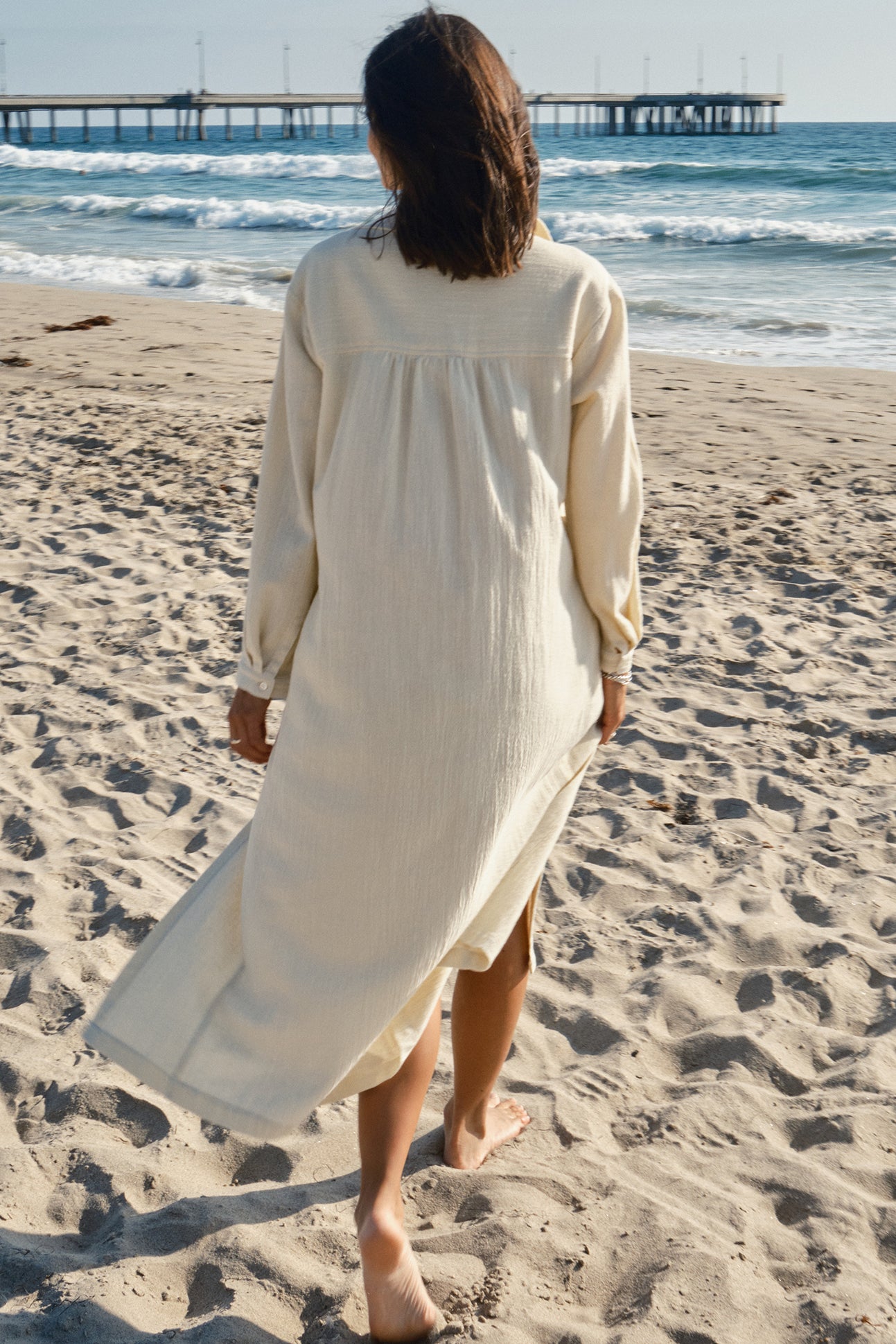 Person wearing a long alabaster dress walking on a sandy beach with a pier in the background.