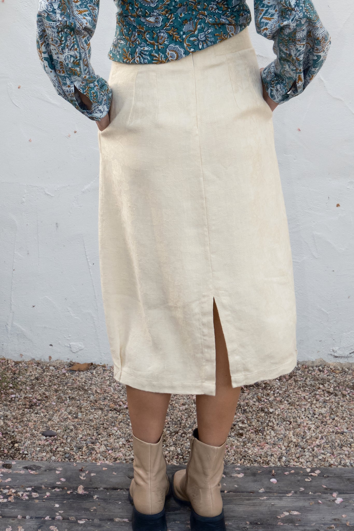 Person wearing a cream skirt with a floral top and boots against a white wall.