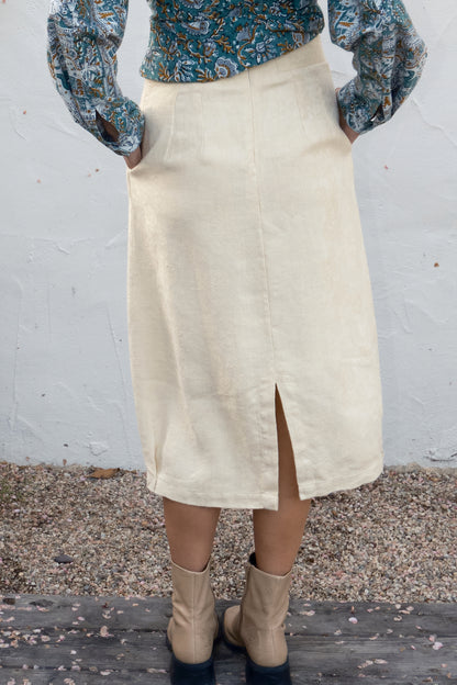 Person wearing a cream skirt with a floral top and boots against a white wall.