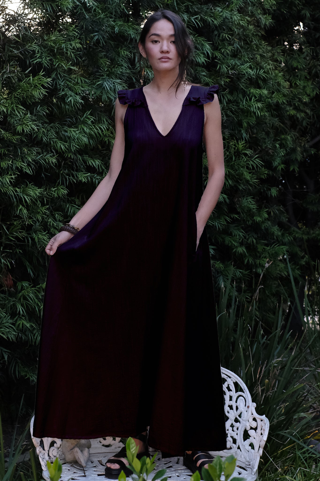 Woman wearing a black dress standing on a white bench with greenery in the background