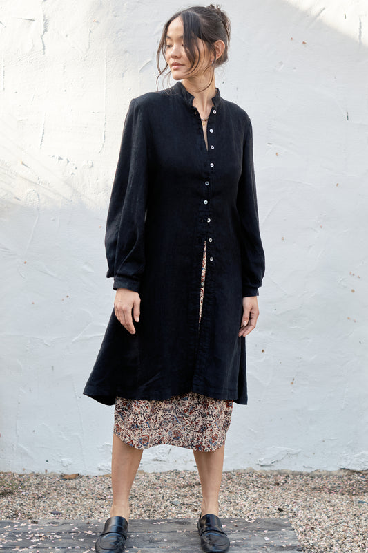 Woman wearing a black coat over a patterned skirt against a white wall.