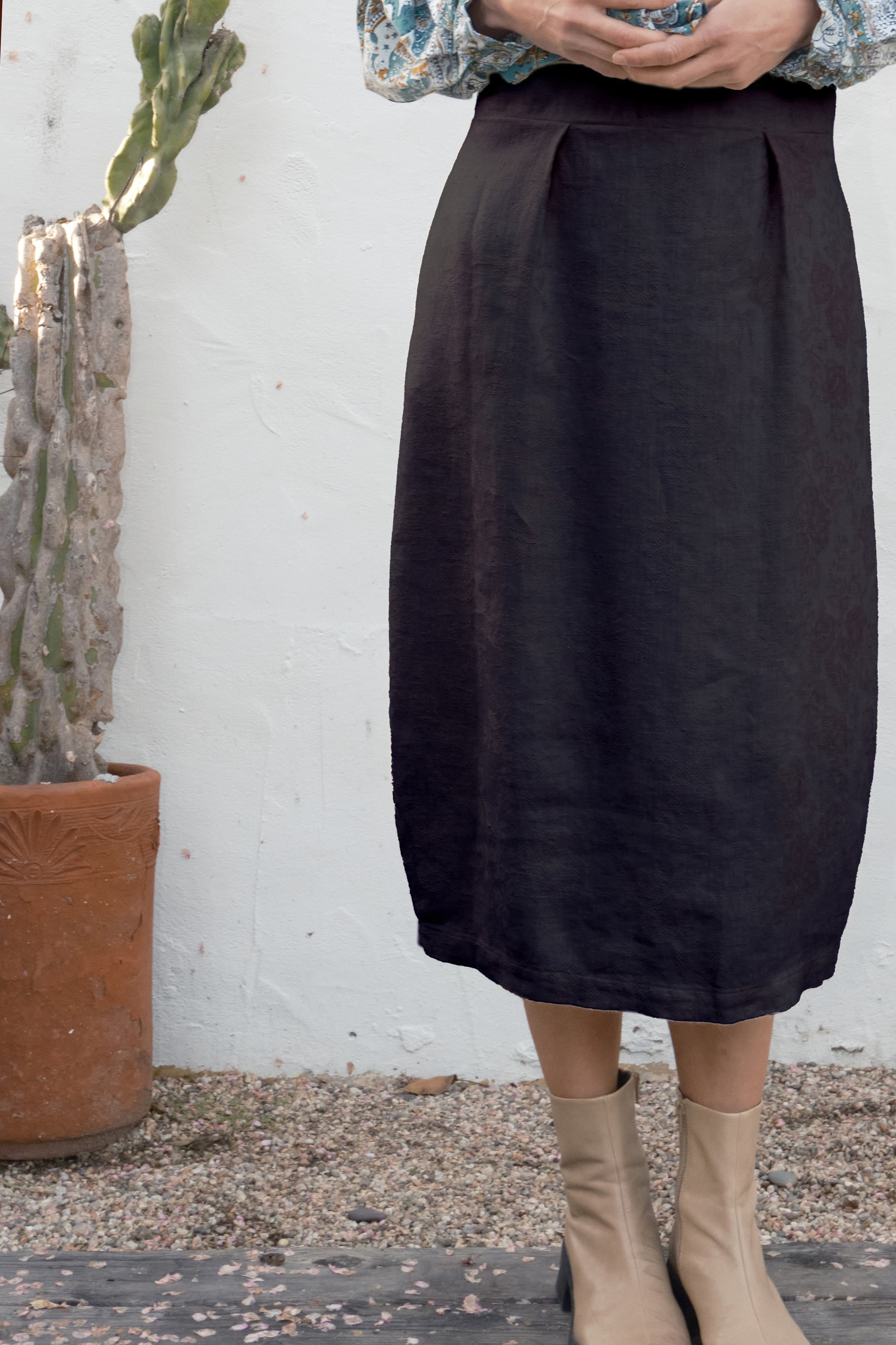 Person wearing a black skirt with beige boots standing against a white wall with a cactus plant.