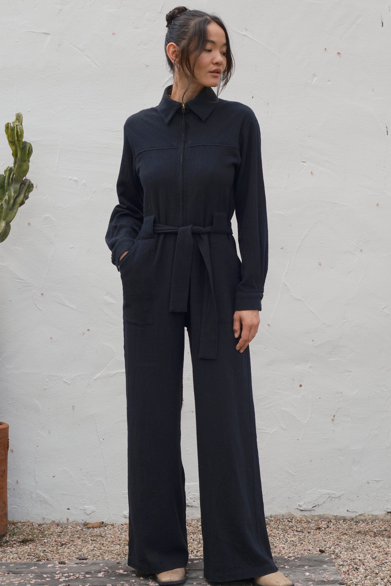 Woman wearing a black jumpsuit standing against a white wall.