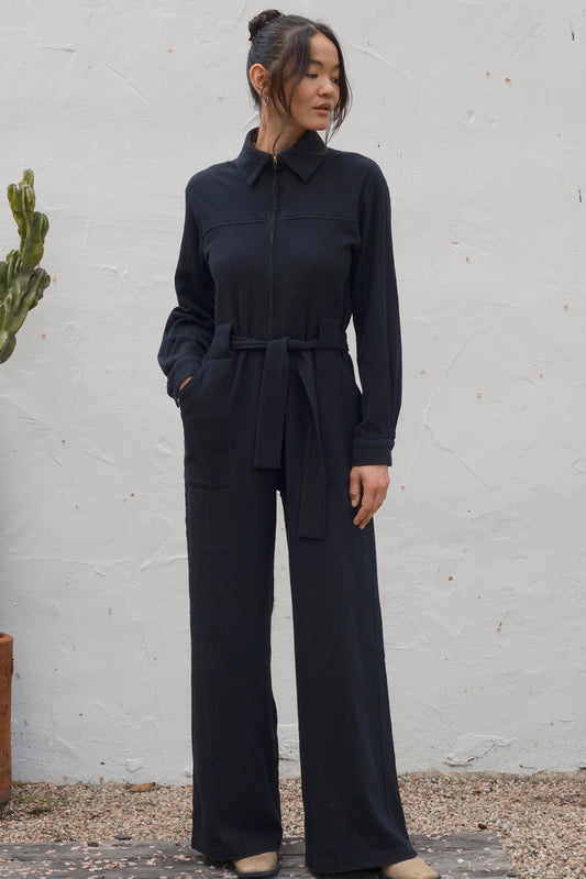 Woman wearing a black jumpsuit standing against a white wall.