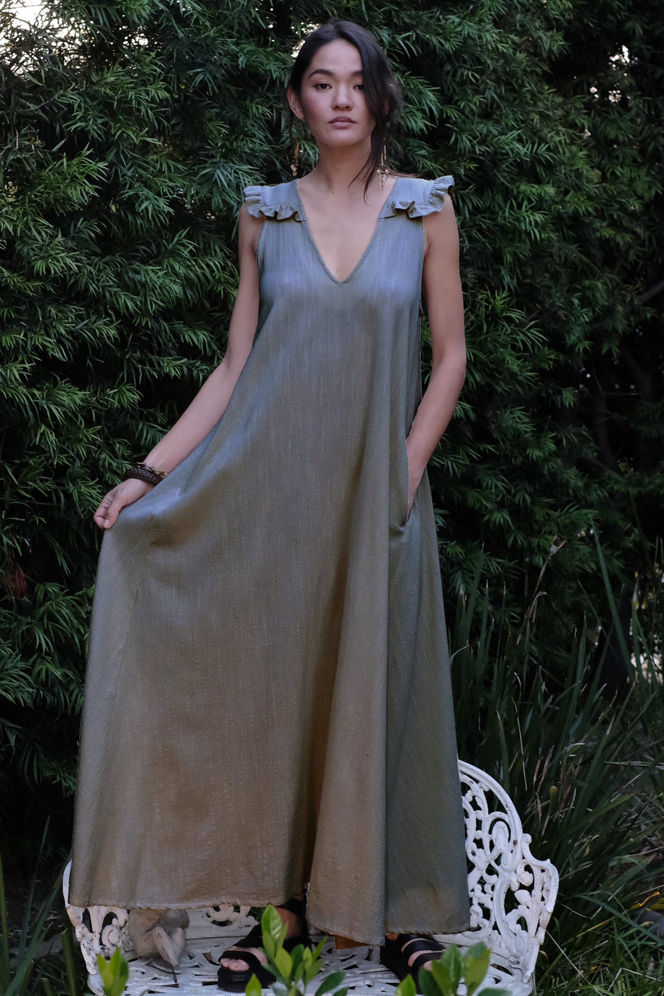 Woman wearing a long, pewter dress standing on a white chair with greenery in the background