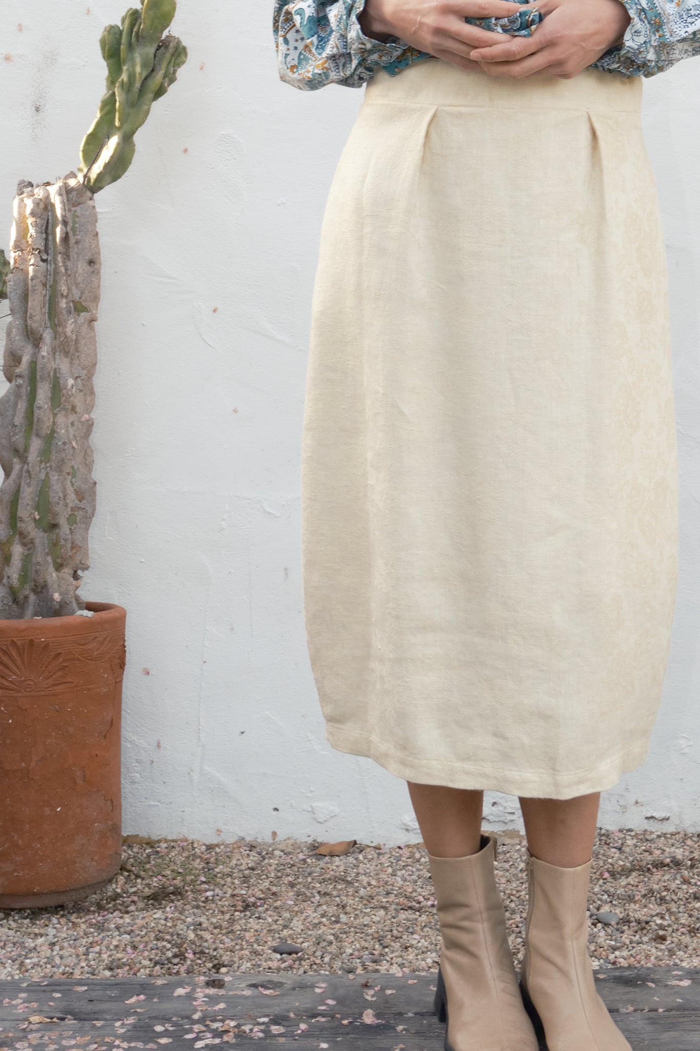 Person wearing a beige skirt and beige boots standing next to a potted cactus against a white wall.