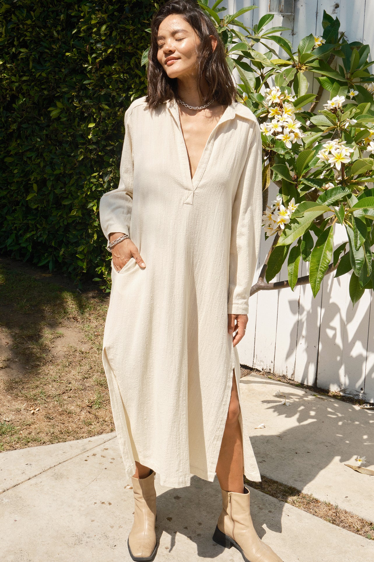 Woman wearing a long beige dress standing outdoors with plants and flowers in the background