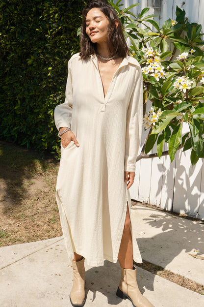 Woman wearing a long beige dress standing outdoors with plants and flowers in the background