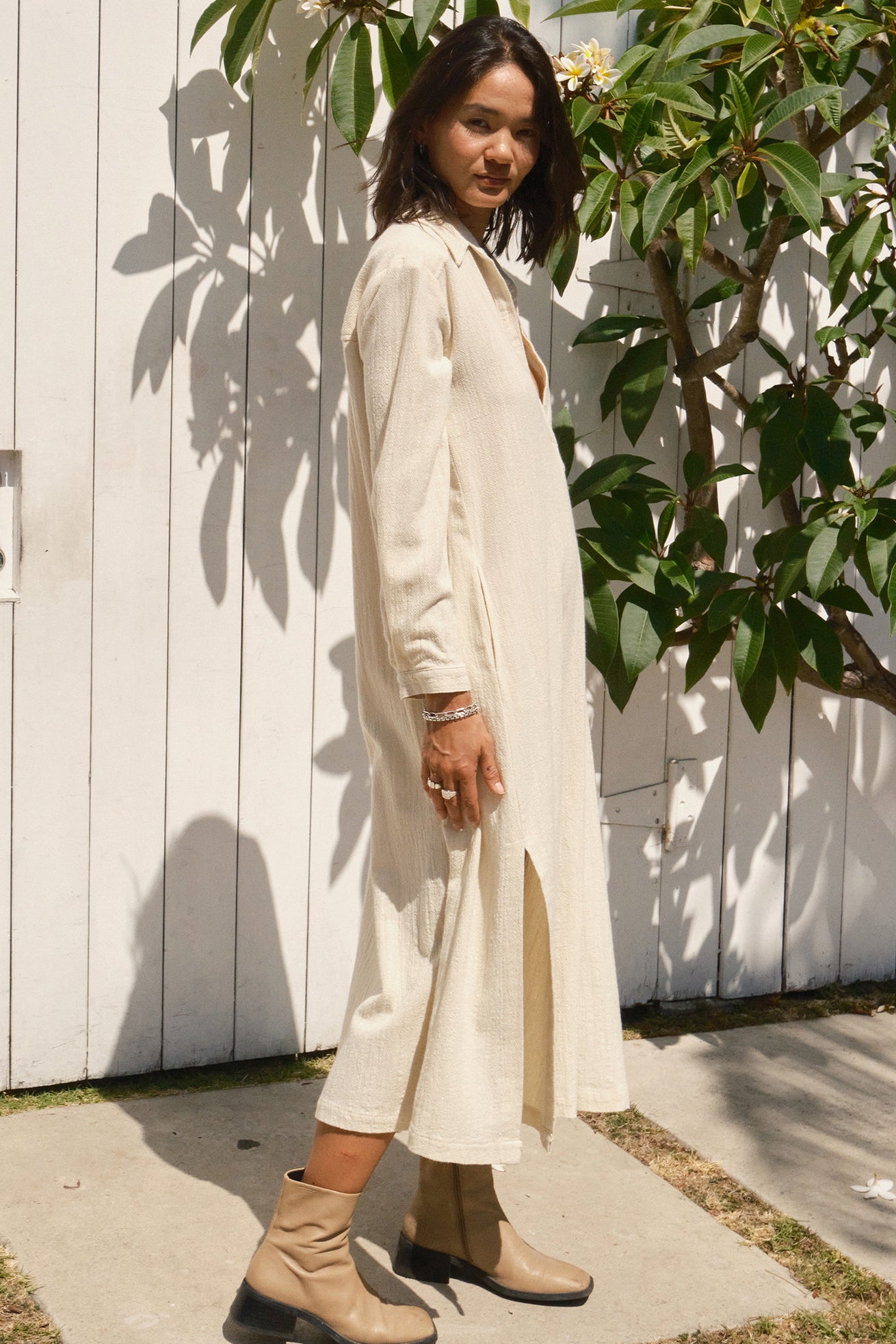 Woman in a long, light-colored dress standing outdoors against a white wall with greenery.