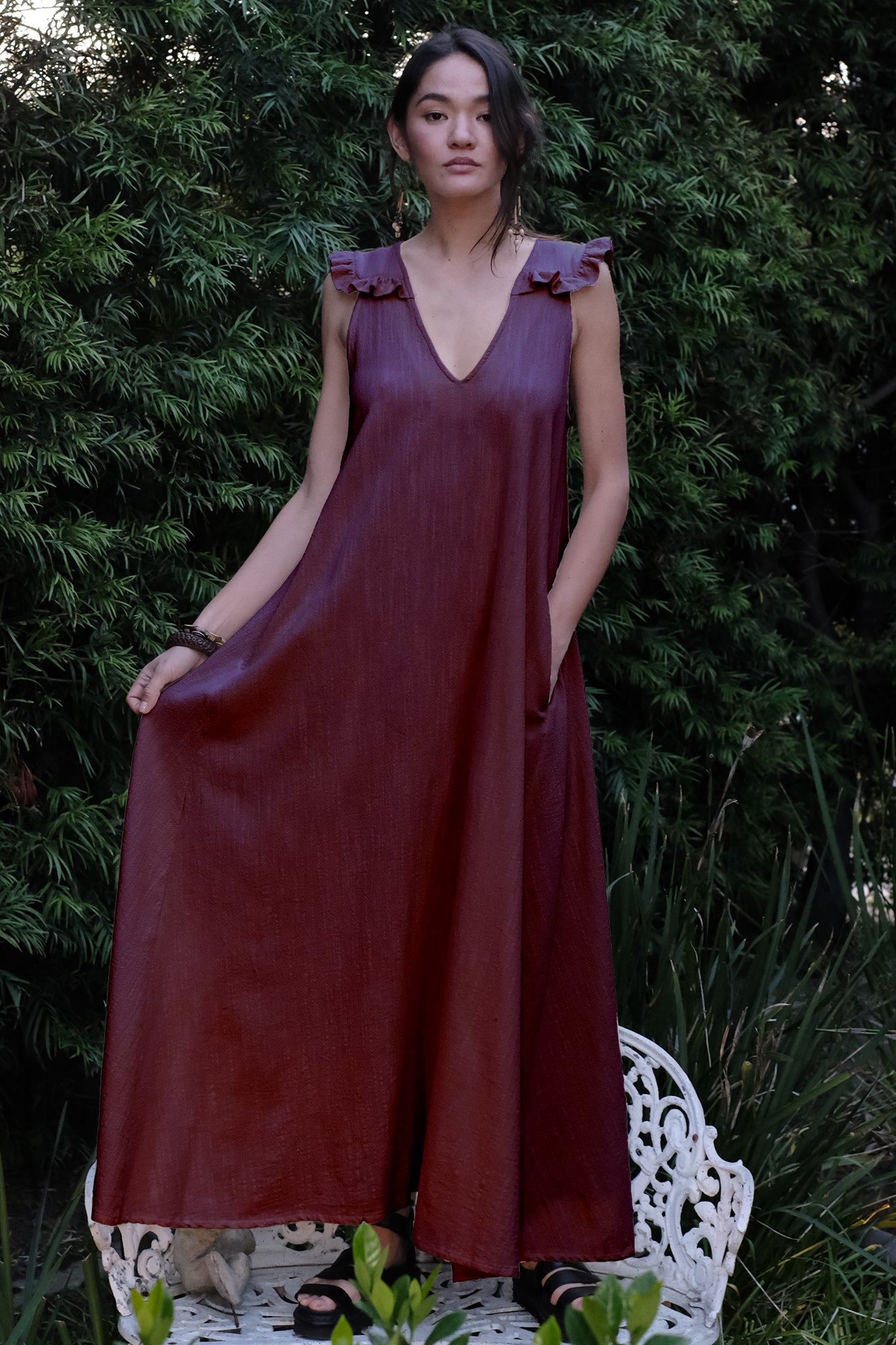 Woman wearing a burgundy dress standing on a white chair with greenery in the background