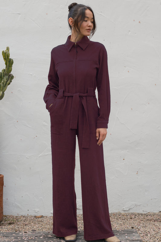 Woman wearing a maroon jumpsuit standing against a white wall.
