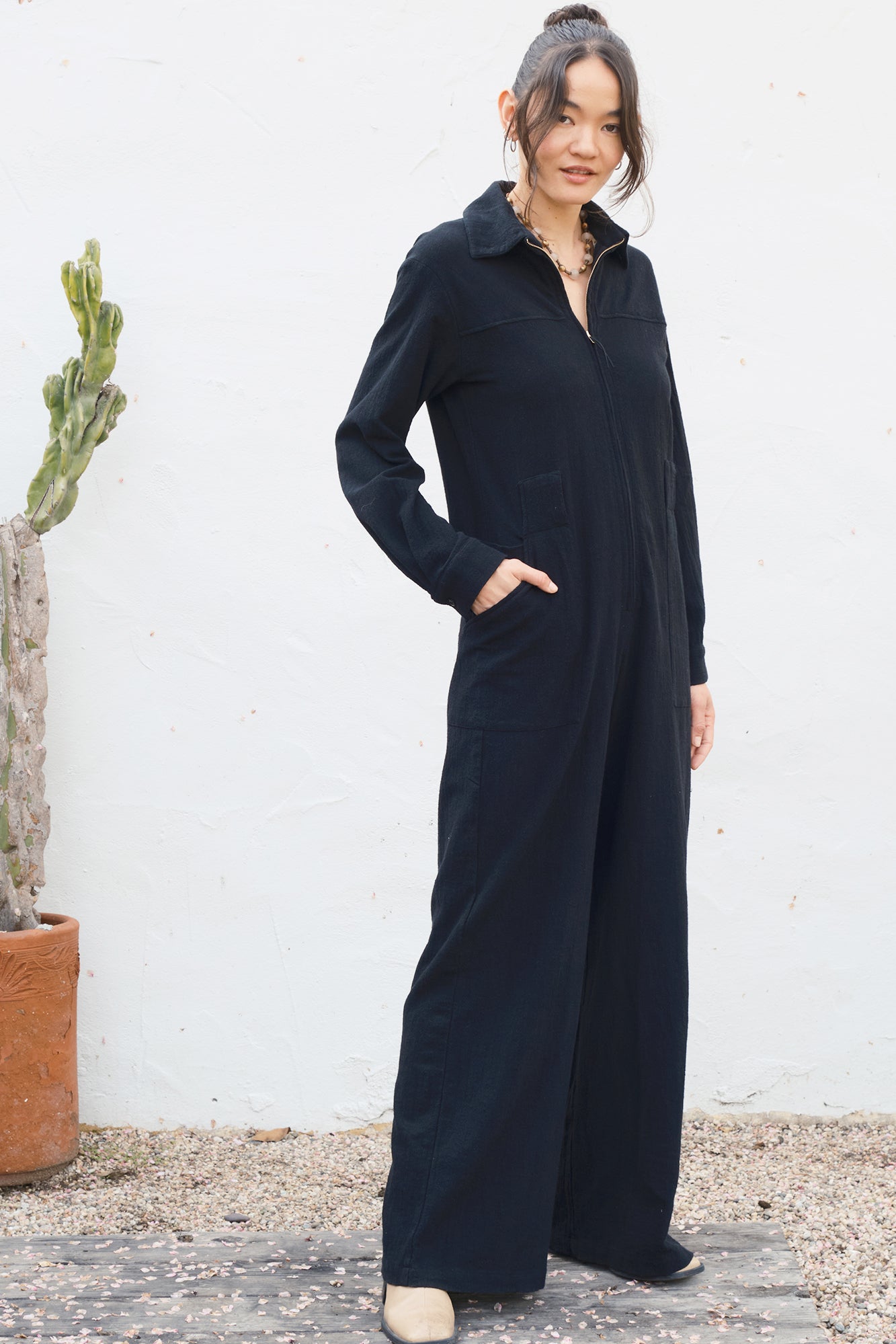 Woman wearing a black jumpsuit standing against a white wall with a cactus plant.