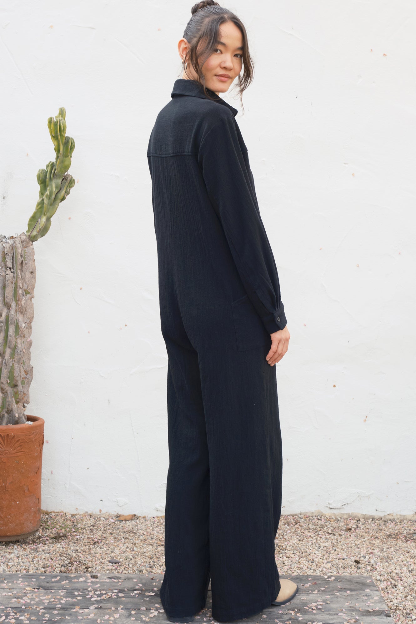 Person wearing a black outfit standing against a white wall with a cactus plant.