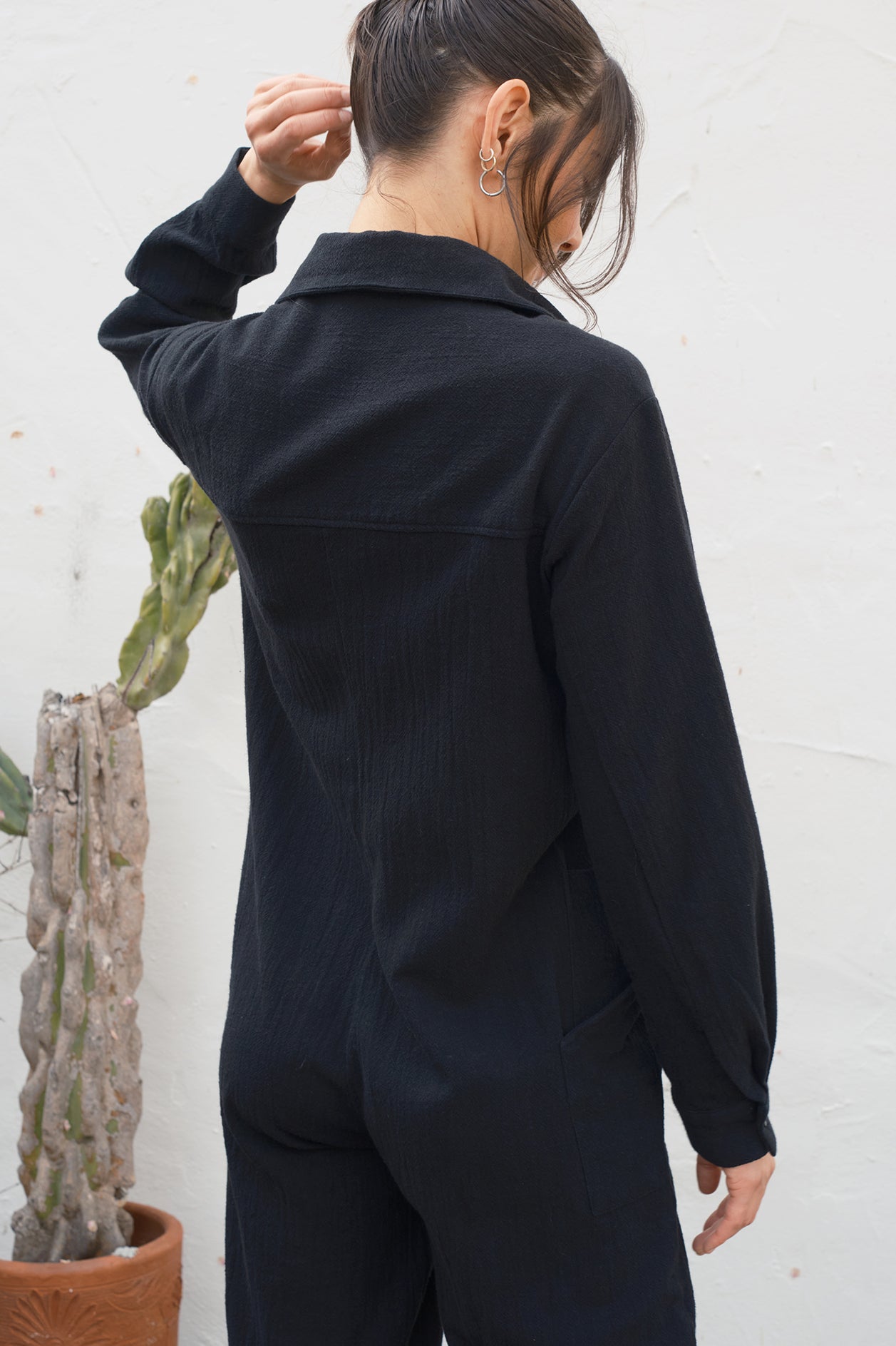 Person wearing a black jumpsuit standing against a white wall with a cactus plant. Back detail