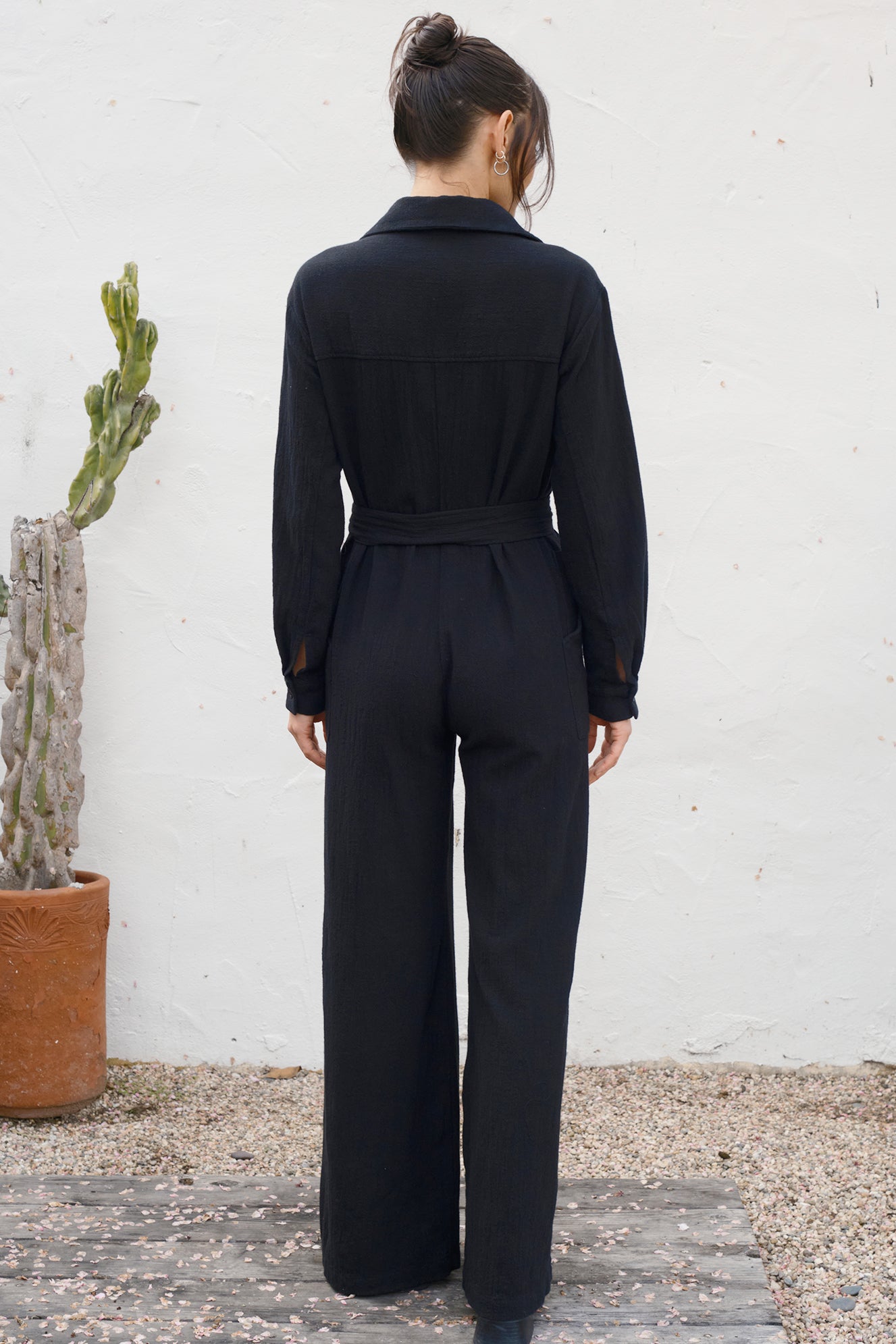 Person wearing a black jumpsuit standing against a white wall with a cactus plant to the left.