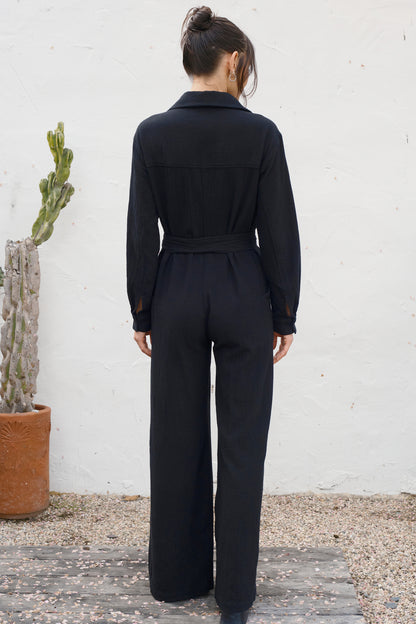 Person wearing a black jumpsuit standing against a white wall with a cactus plant to the left.