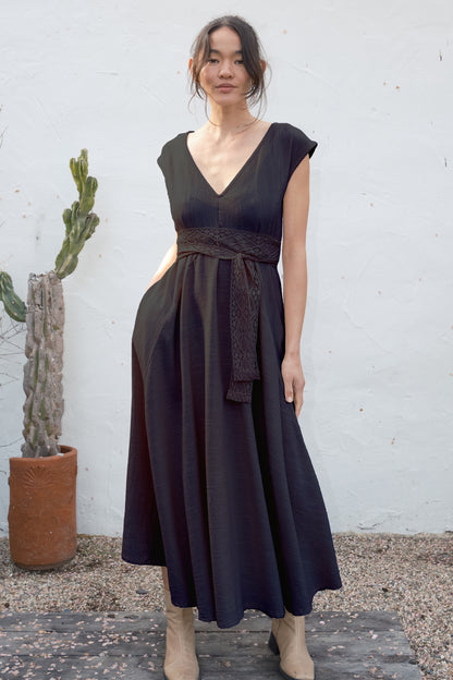 Woman wearing a black dress with a belt, standing against a white wall with a cactus plant.