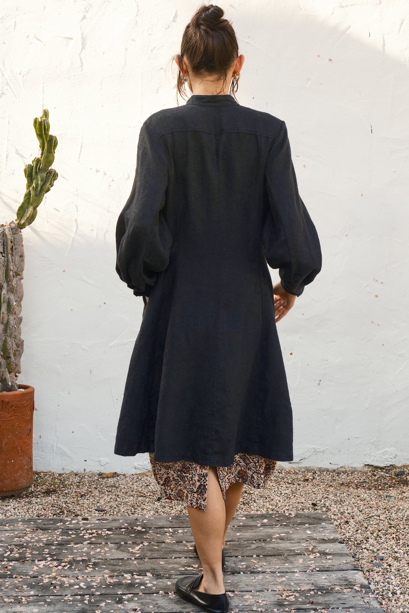 Person wearing a black coat over a floral hem, standing against a white wall. Back view