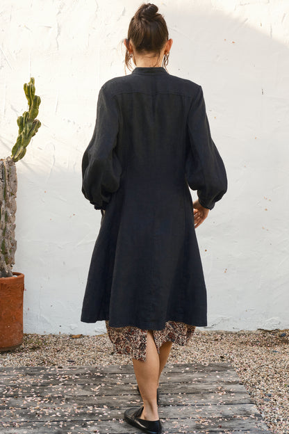 Person wearing a black coat over a floral hem, standing against a white wall. Back view