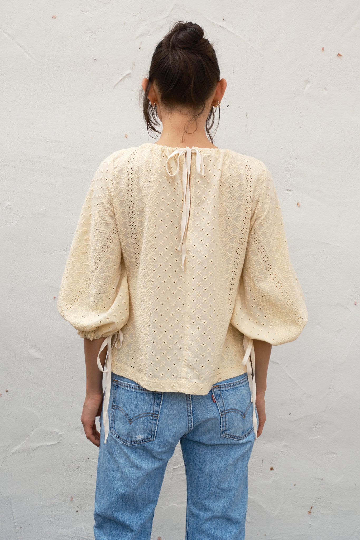 Person wearing a cream blouse with lace details and blue jeans against a light gray background