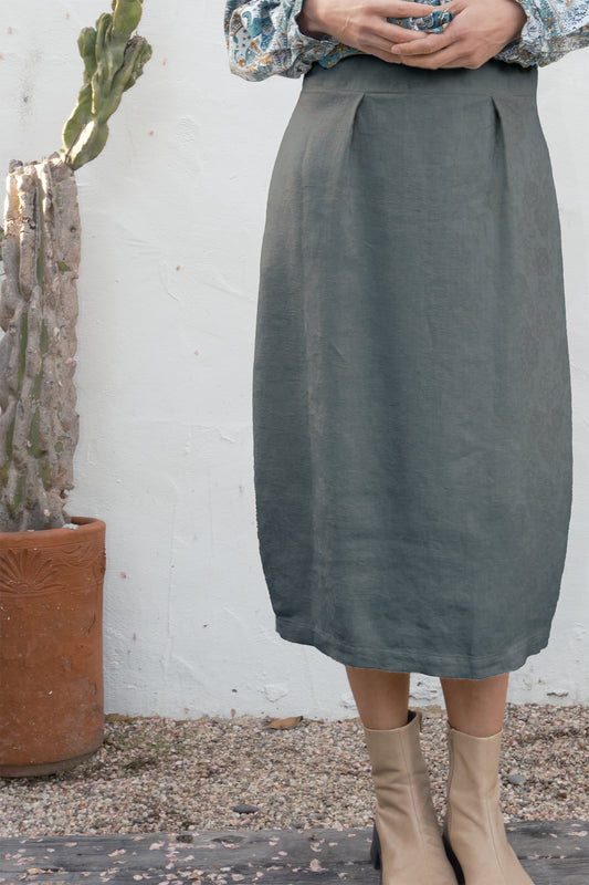 Person wearing a gray skirt standing next to a cactus plant against a white wall.