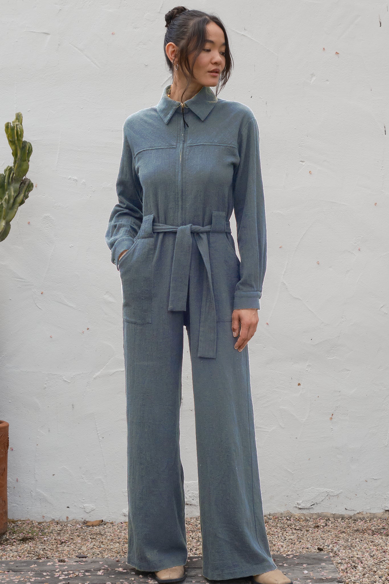 Woman wearing a gray jumpsuit standing against a white wall.