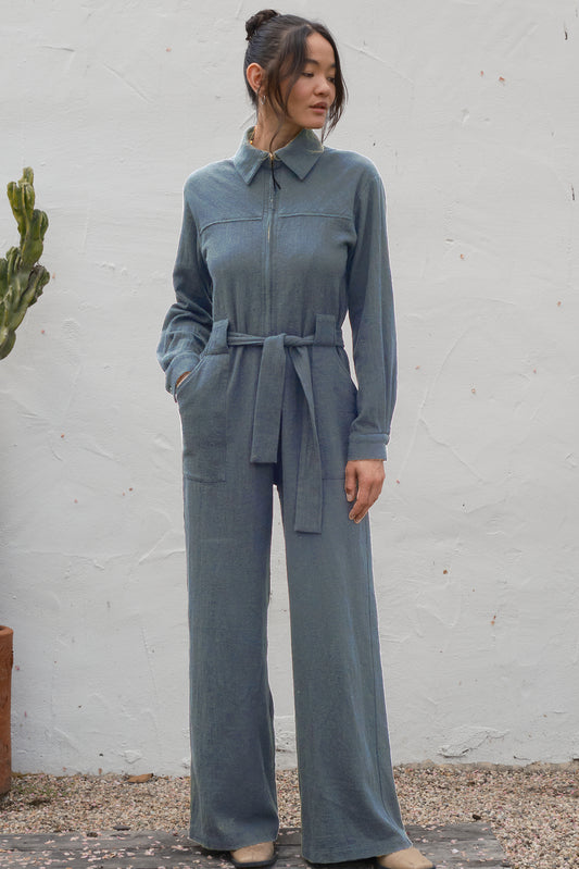 Woman wearing a gray jumpsuit standing against a white wall.