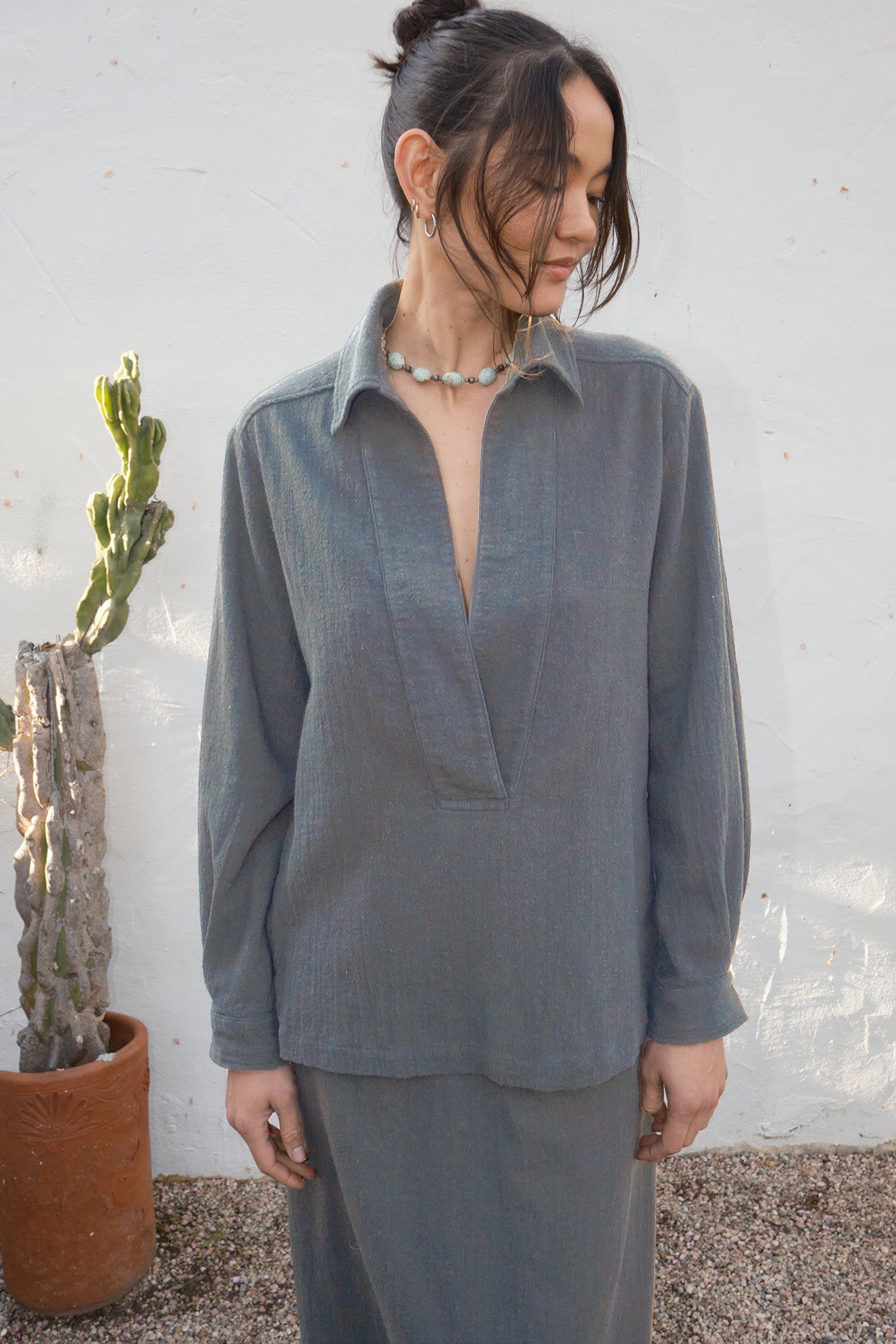 Woman wearing a gray long-sleeve top and skirt  standing next to a potted plant against a white wall.