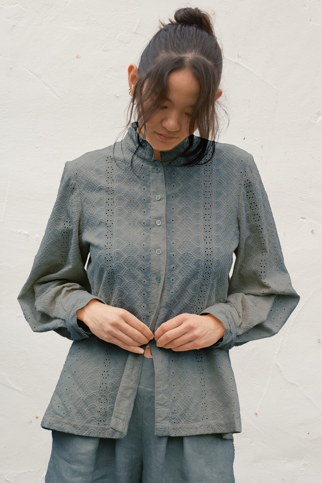 Woman wearing a gray blouse with intricate patterns against a white wall.