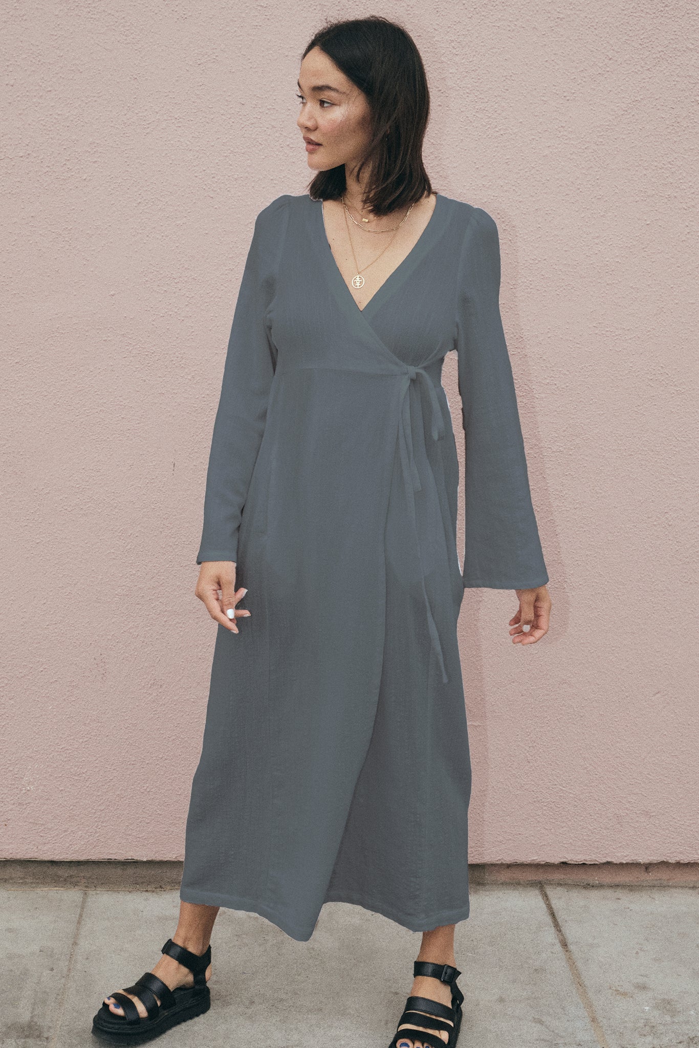 Woman wearing a gray wrap dress against a pink wall.