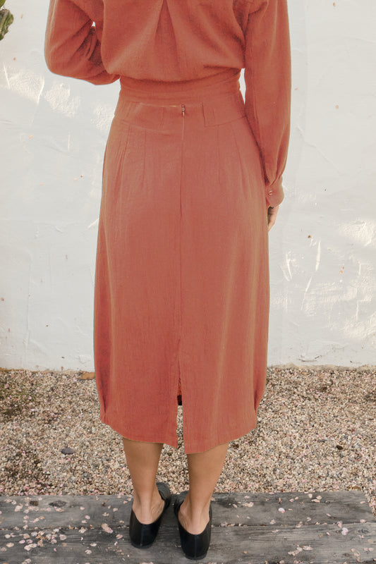 Person wearing a rust-colored dress standing on a gravel surface with a white wall background