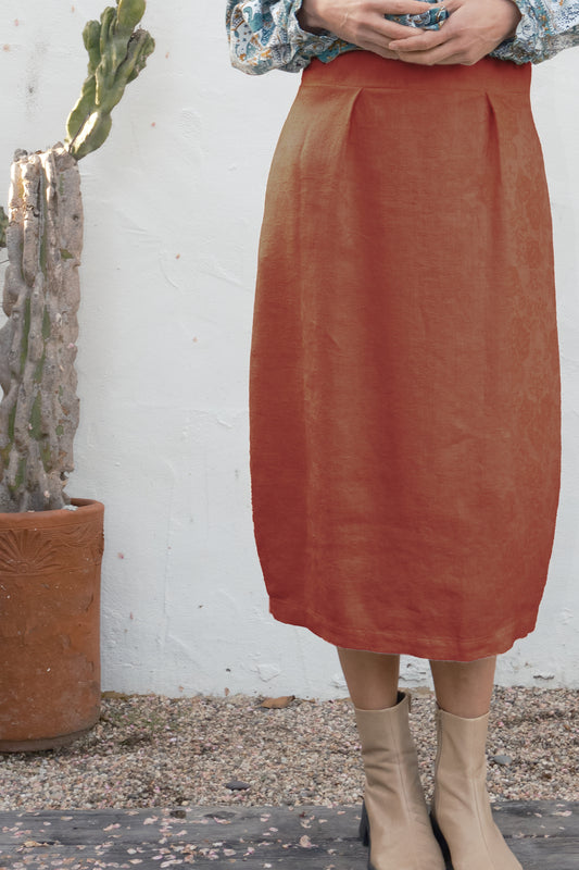 Person wearing a rust-colored skirt standing next to a cactus plant against a white wall.