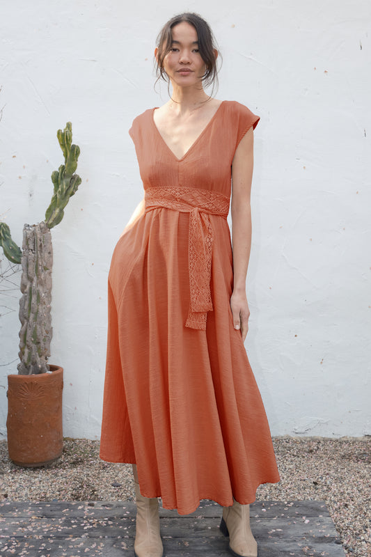 Woman wearing a rust-colored dress standing against a white wall with a cactus plant.