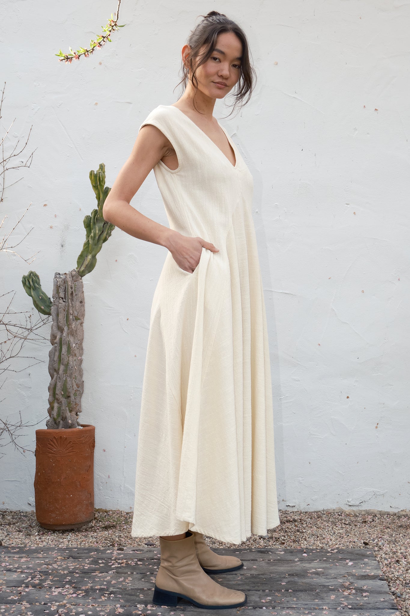 Woman wearing an alabaster  dress standing against a white wall with a plant in the background