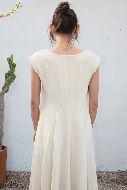 Person wearing an alabaster dress standing against a light gray wall with a plant in the background.