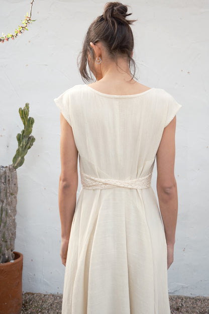 Woman wearing an alabaster dress against a plain wall with a plant in the background