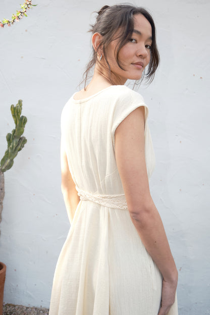 Woman wearing an alabaster  dress against a light gray wall with a plant in the background